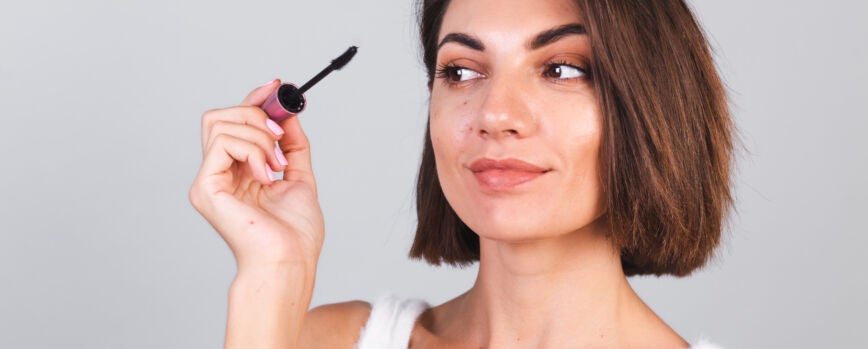 Linda mulher com maquiagem segurando pincel de máscara negra sobre parede cinza, conceito de beleza