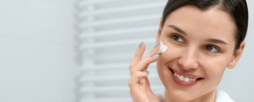 Mulher sorridente em bata branca com creme facial no banheiro.