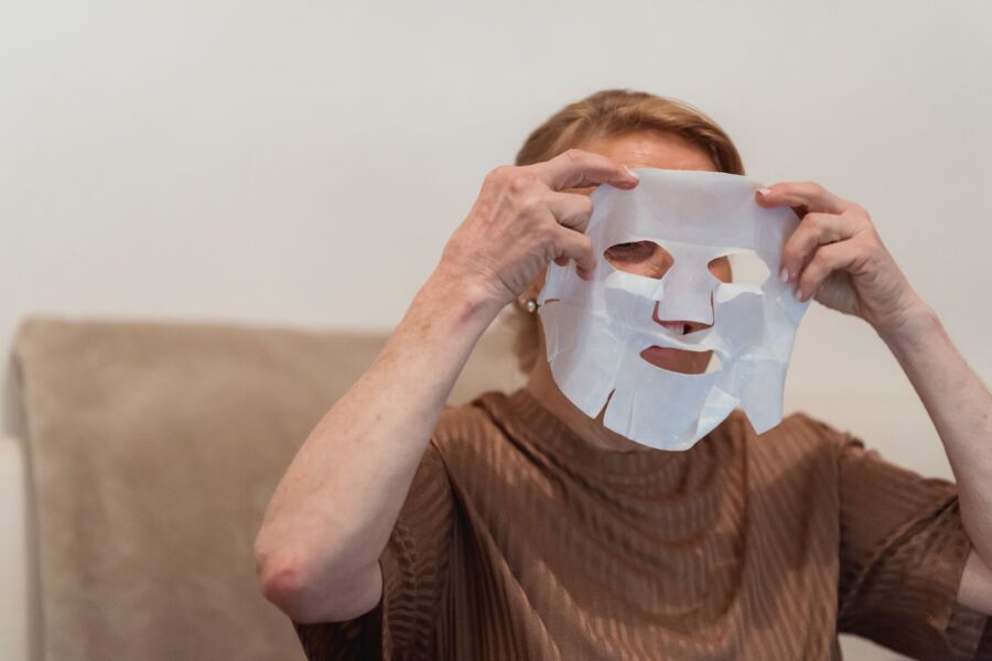 Mulher idosa aplicando uma máscara de tecido para cuidados com a pele e relaxamento em ambientes internos.