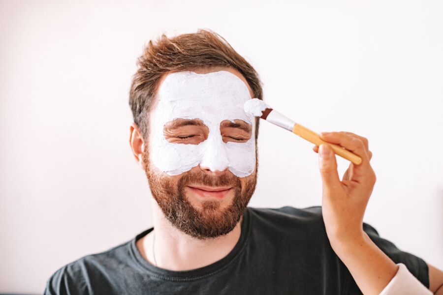 Um homem com barba desfrutando de um tratamento de spa em casa com uma máscara aplicada com um pincel.