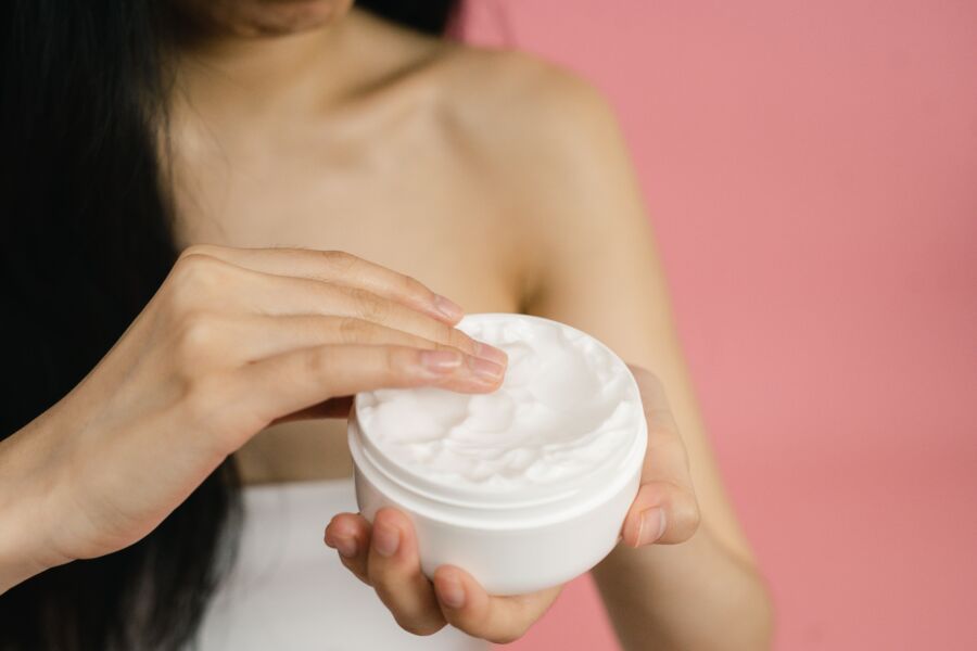 Close-up de uma mulher aplicando creme branco de um frasco sobre um fundo rosado.