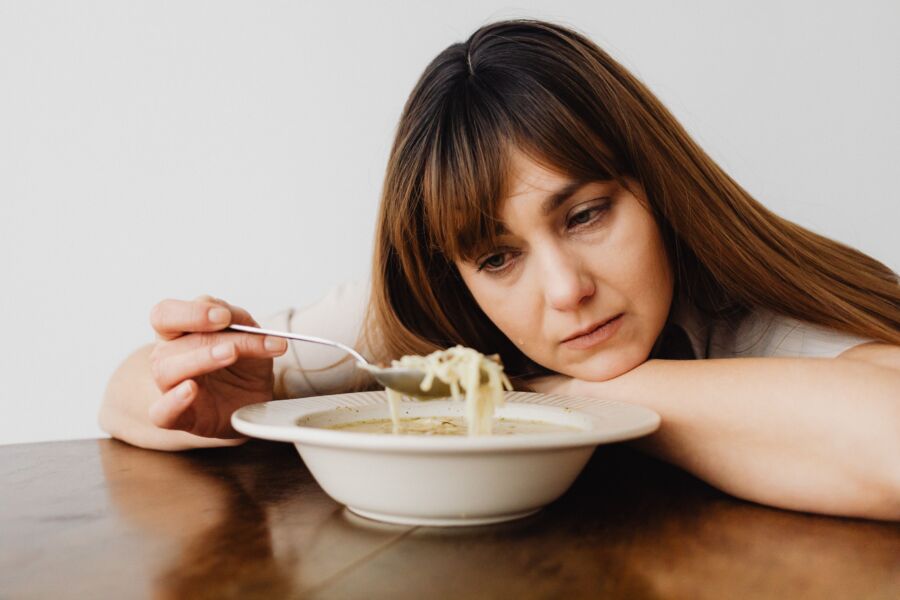 Uma mulher sentada à mesa olhando pensativamente para uma tigela de sopa de macarrão, evocando um sentido de contemplação.