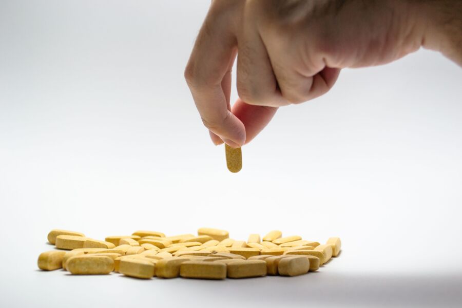 Close de uma mão pegando uma pílula amarela de uma pilha de comprimidos sobre uma superfície branca.