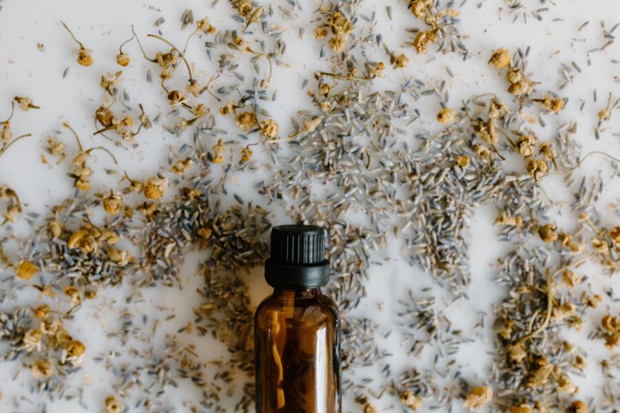 Close-up de uma garrafa de óleo essencial cercada por lavanda seca e camomila sobre uma superfície branca.