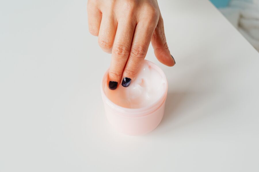 Close-up de uma mão com as unhas pintadas mergulhando em um frasco de creme facial, representando o autocuidado.