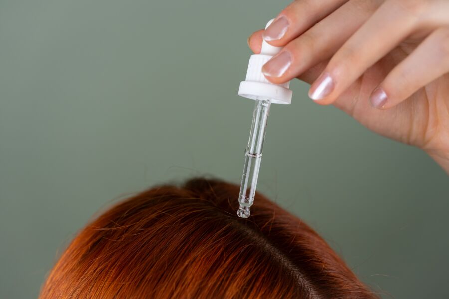 Primeiro plano da aplicação de sérum nas raízes com um conta-gotas de precisão sobre o cabelo vermelho, mostrando a rotina de cuidado do cabelo.