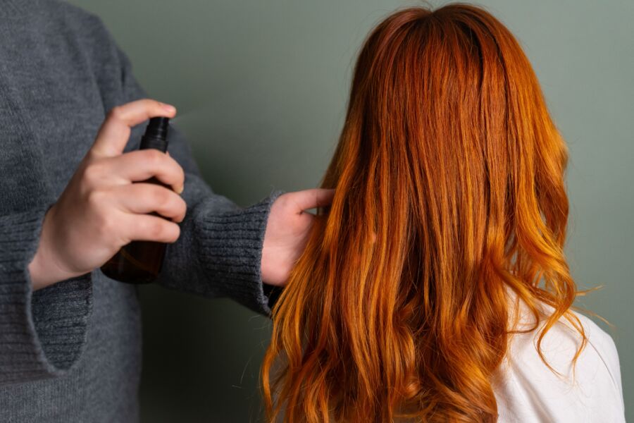 Primeiro plano de cabeleireiro pulverizando produto capilar sobre cabelo longo de cor vermelha.