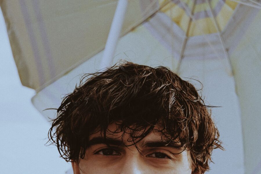 Retrato de meia face de um jovem com cabelo bagunçado sob um guarda-sol de praia.