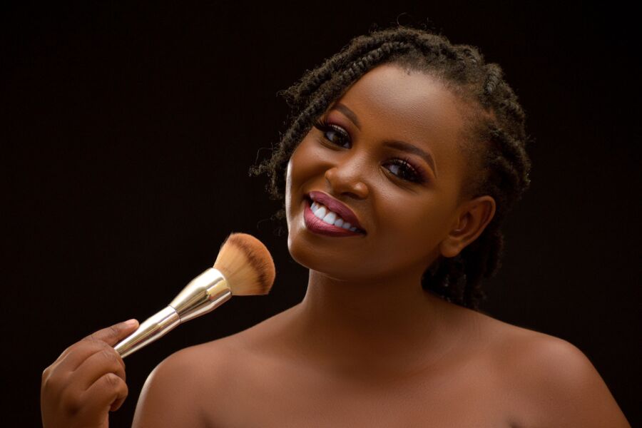 Uma bela mulher sorridente com cabelos trançados segurando um pincel de maquiagem contra um fundo preto.