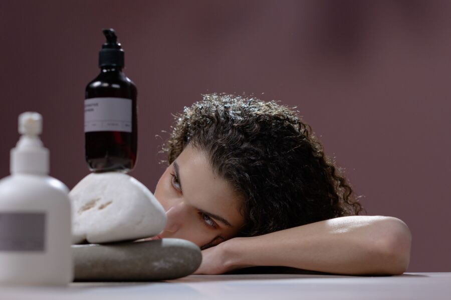 Uma mulher com cabelo encaracolado se inclina sobre uma mesa segurando garrafas de cosméticos em cima de pedras empilhadas.
