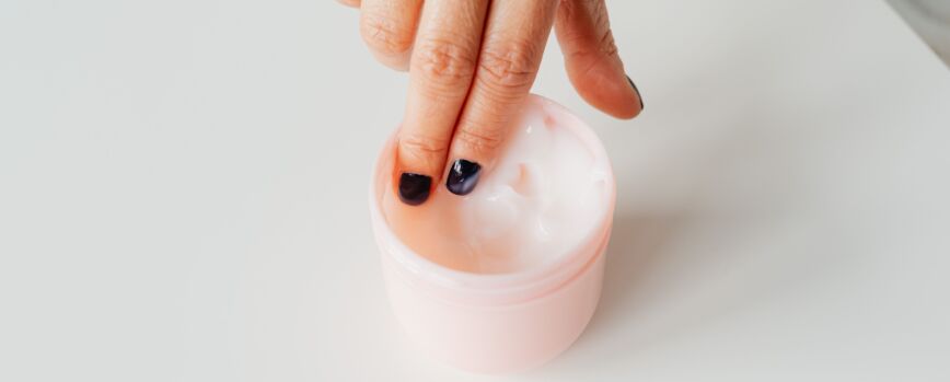 Close de uma mão com unhas pintadas mergulhando num pote de creme facial, representando o autocuidado.