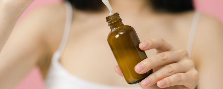 Close de uma mulher usando um soro cosmético de um frasco conta-gotas contra um fundo rosa.